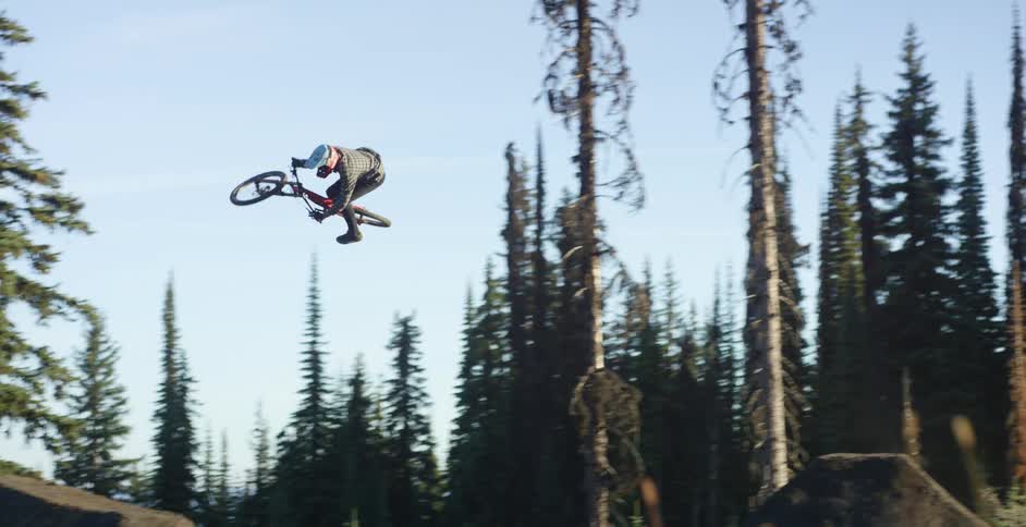 Man on a ride trick bike rides over a large dirt slope and performs a trick in the air in a forest at daytime