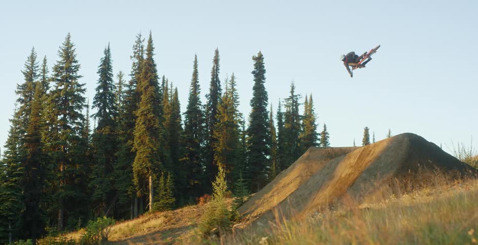 A bicyclist leaps off a dirt mound and spins in the air near a forest