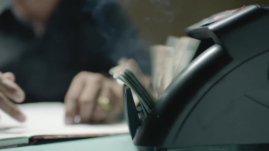 A man sits at a desk and presses a button on a machine that counts cash as he looks at a ledger book