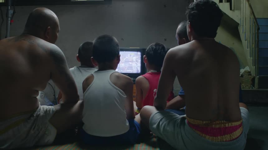Three young boys sit on the floor with older male family members and watch a boxing match on an old television