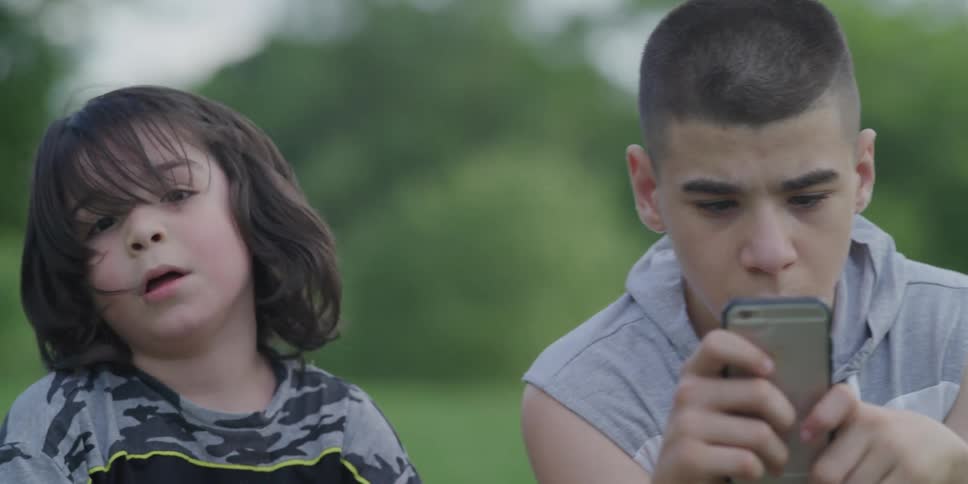 A teenager on an iphone and a younger boy next to him playing with a soccer ball sitting in the grass outside