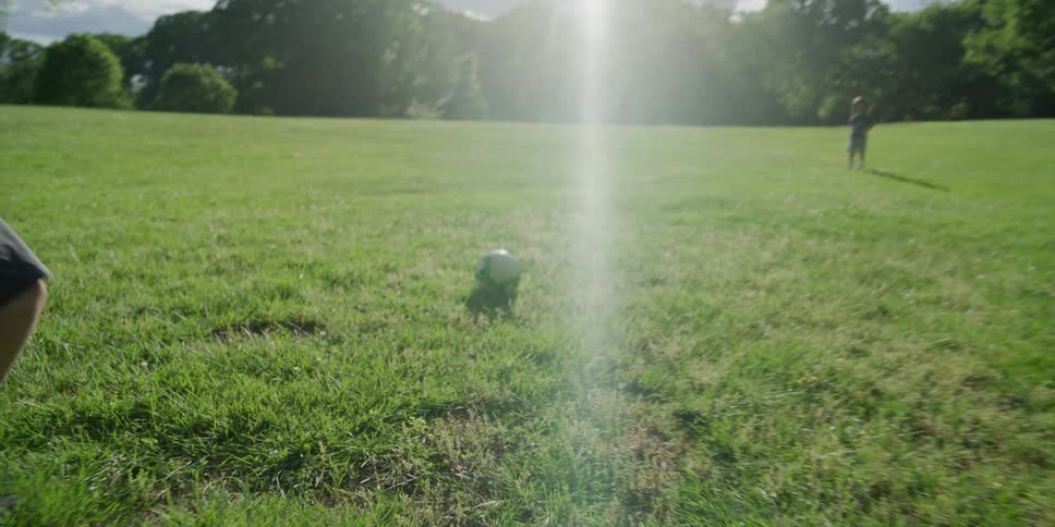 Two boys walking and running on a grassy field kicking a soccer ball with trees the sky and clouds in the background