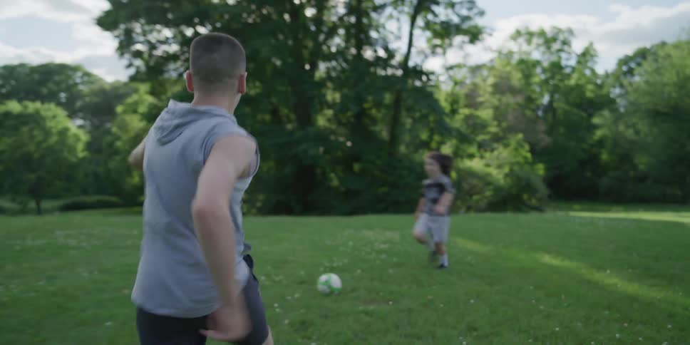 Two boys running in a grassy field as one kicks a soccer ball with trees the sky and clouds in the background