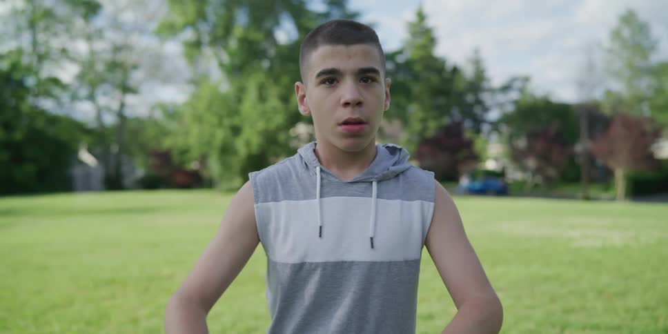 A hispanic teenage boy in a sleeveless hoodie shirt is posing for the camera in an open field in front of houses