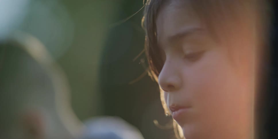 A boy sits outdoors to another boy with sunlight visible