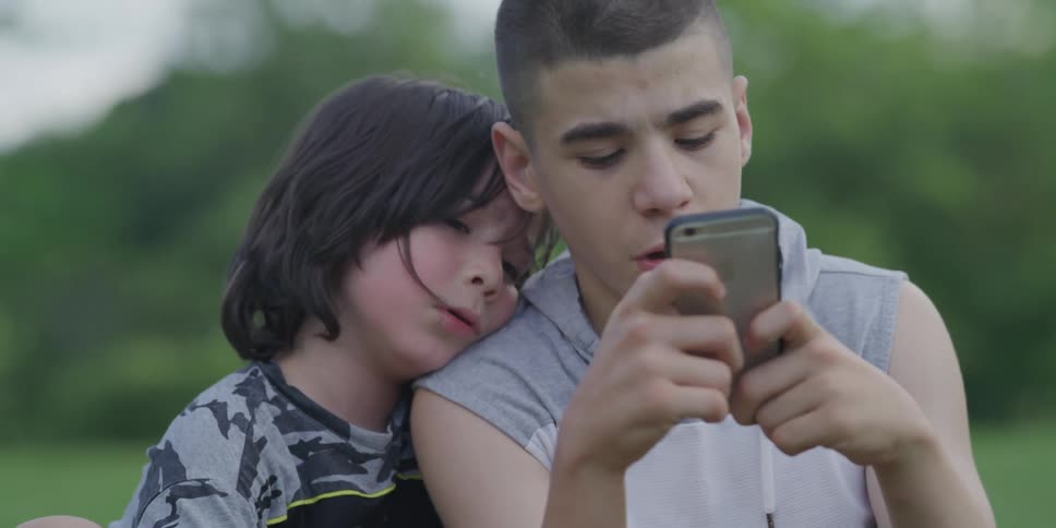 Brothers sitting outside on a lawn while the older one uses a smartphone and the little one watches from his shoulder