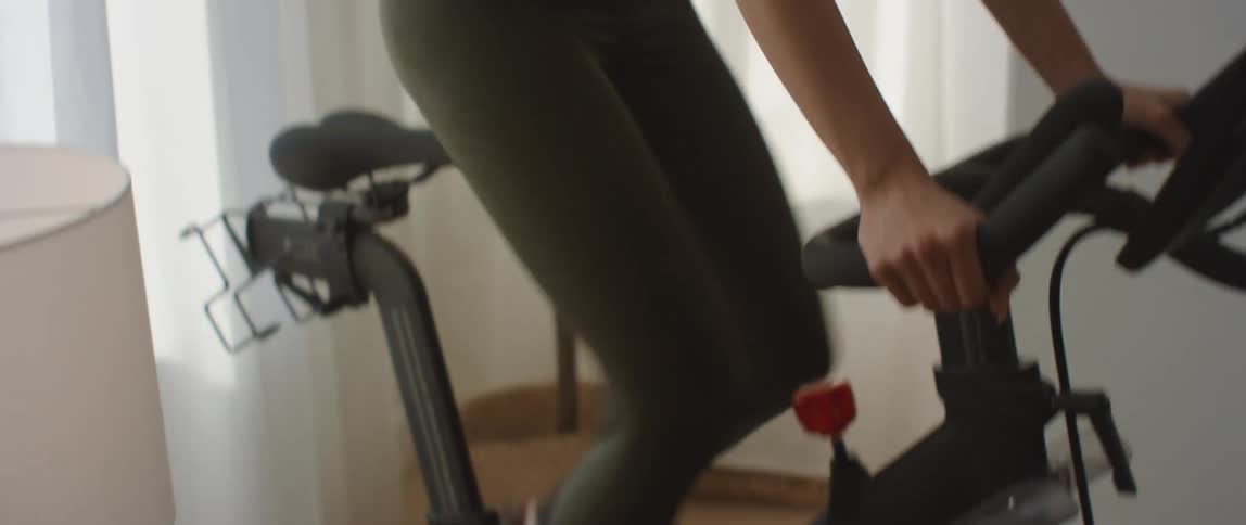 A young woman stands while pedaling on an exercise bike in house during the day