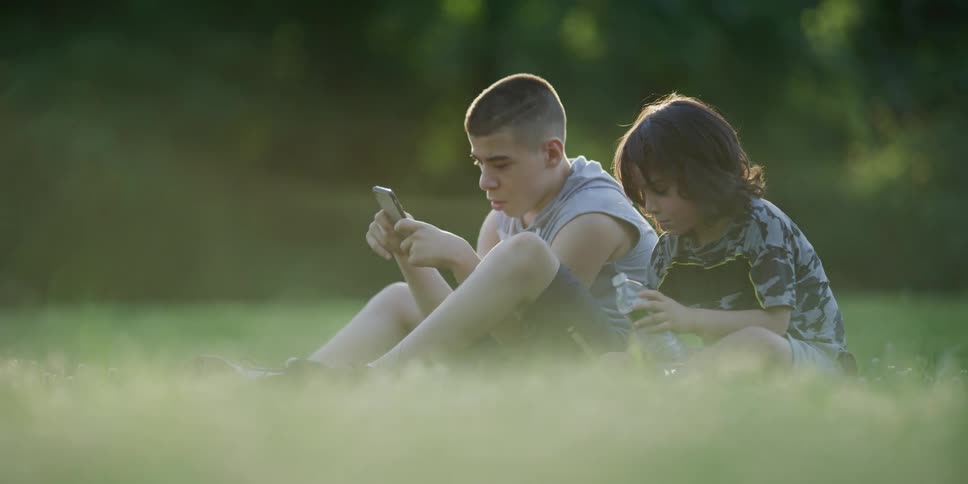 A boy plays on his cell phone while sitting next to a younger boy on the lawn
