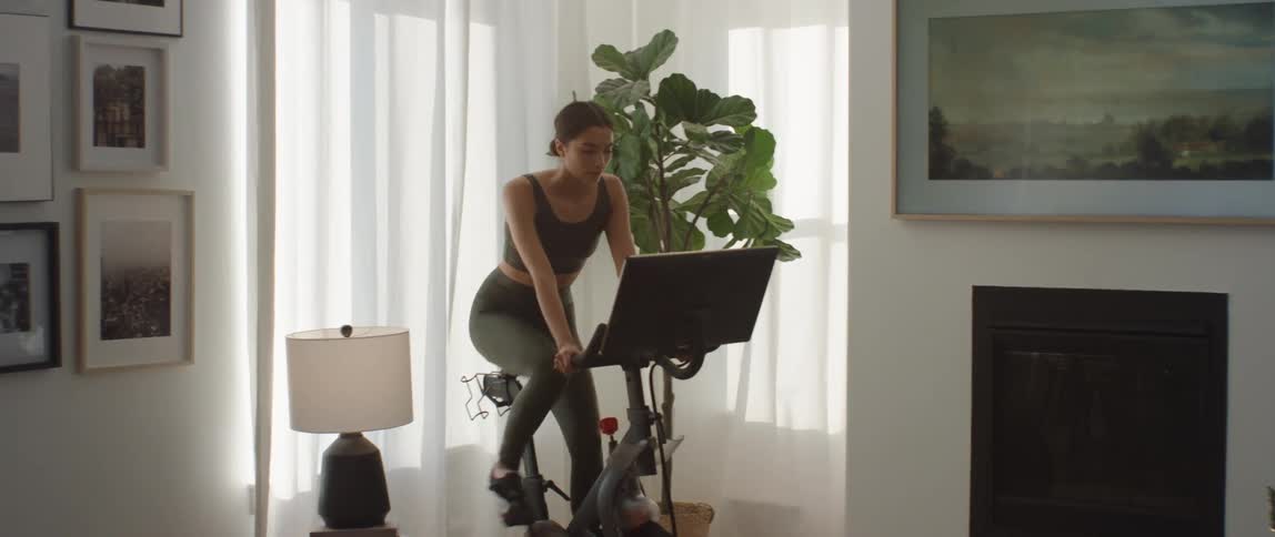 Woman pedaling on a pelaton workout bike in a living room at daytime