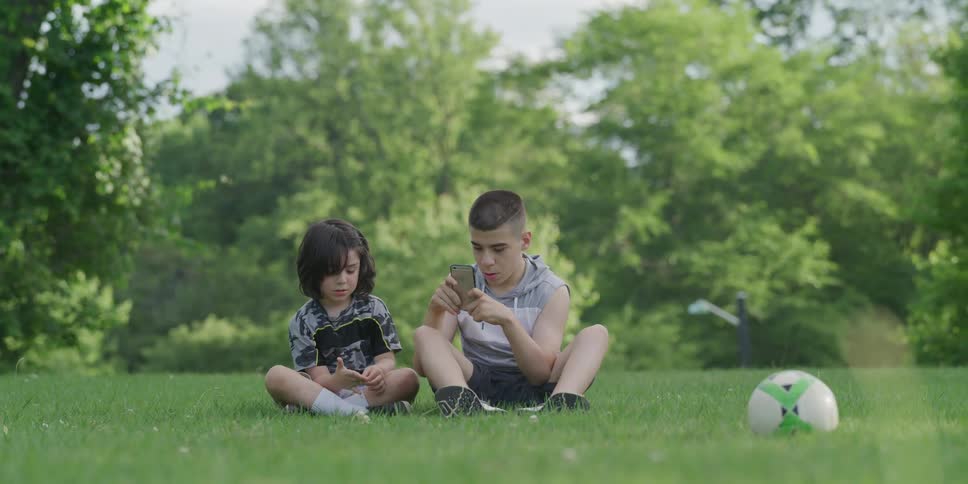 A boy sits on the grass outdoors with another boy who is beside him texting on his cell phone