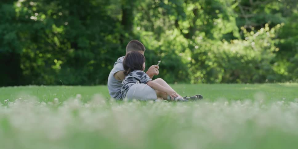 A boy sits on a lawn next to his brother as he plays on his cell phone