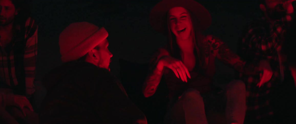 Woman sitting outdoors at nighttime with friends tips her hat and laughs