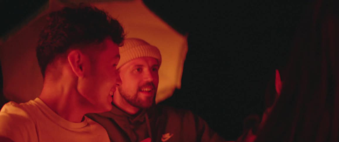 A man shakes his friends shoulders as they sit around a campfire at night