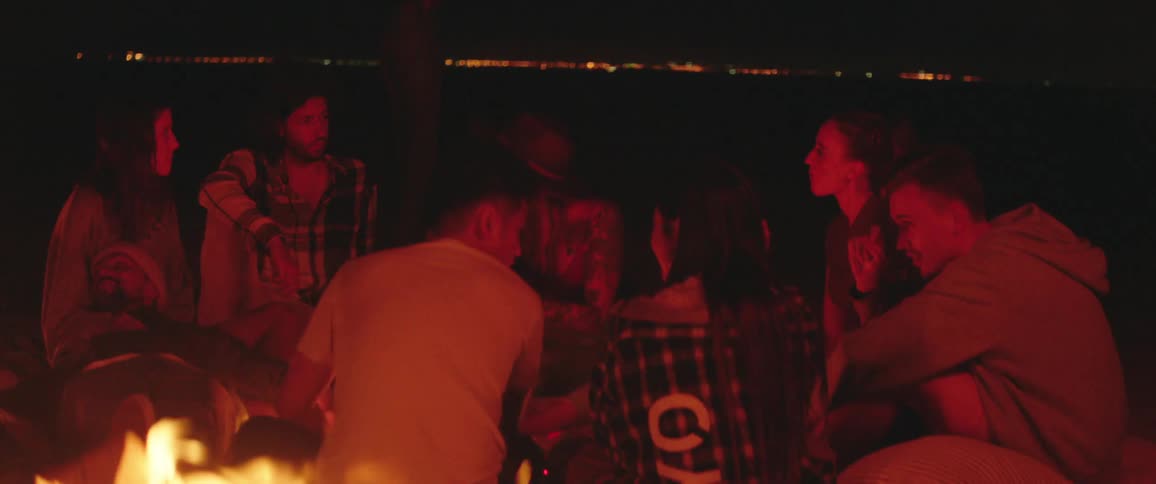 A group of people chat while sitting in a circle near a campfire at night