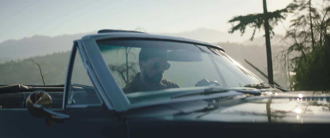 A man sits in a convertible car in a rural setting and then drives away