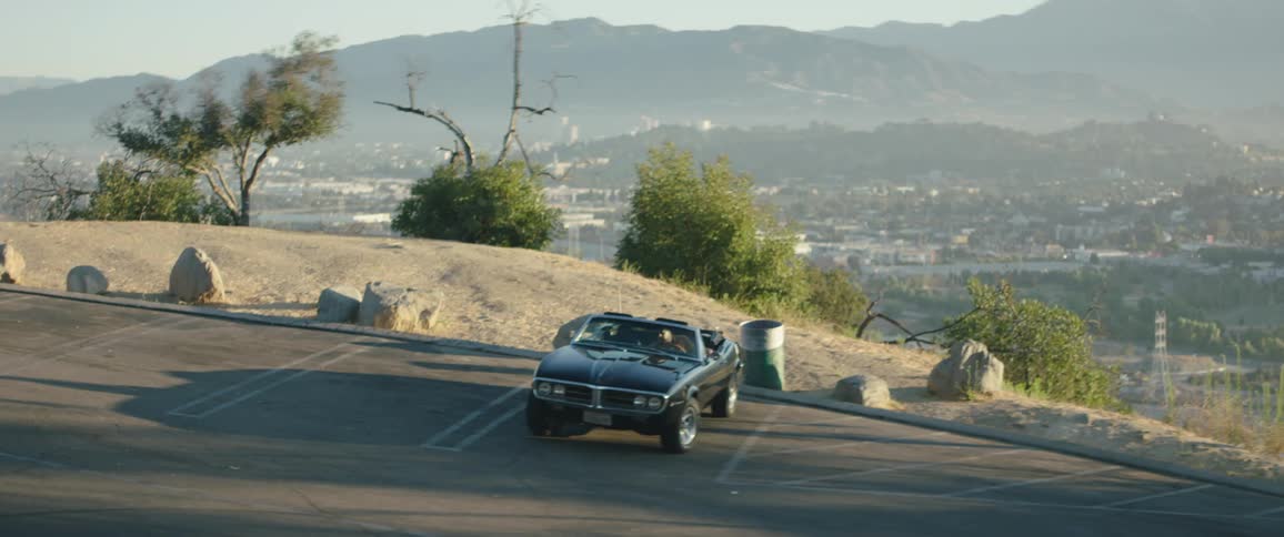 A man walks through a parking lot on a hill overlooking a city and enters the drivers seat of a convertible