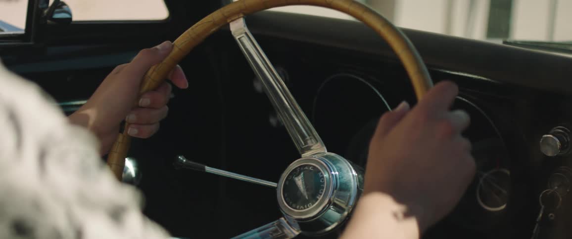 A woman sits in a vintage car with her hands on the steering wheel