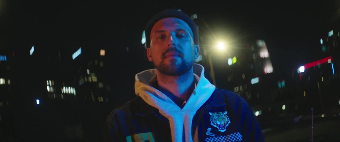 A man faces the camera wearing a hoodie sweatshirt and jacket while standing outdoors at night