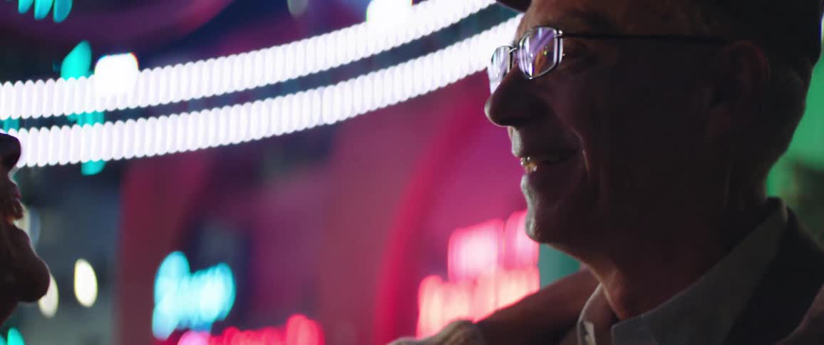 An elderly man and woman dance together on a colorful carnival ground outdoors at night