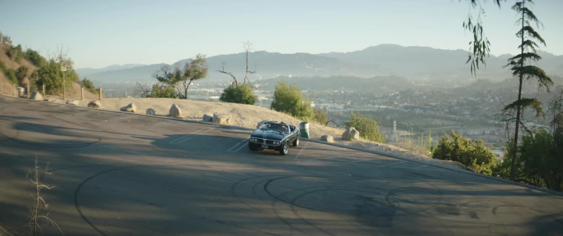 A person walks across a parking lot on a hill overlooking a city and climbs into the drivers seat of a sports car