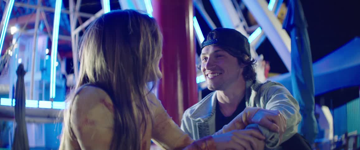 A man and woman chat while sitting together under colorful lights on a fairground at night