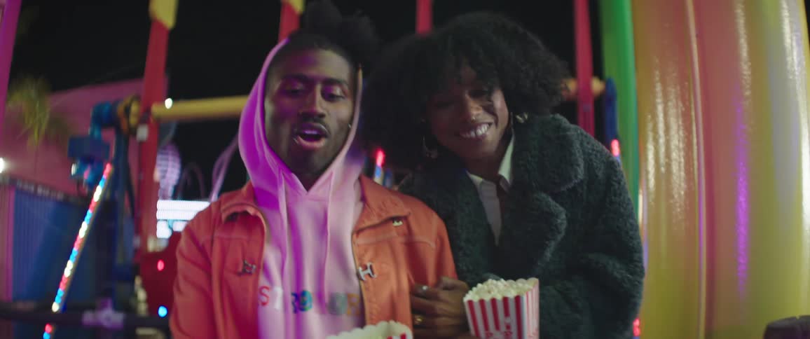 A man and woman carrying popcorn walk through a carnival laughing