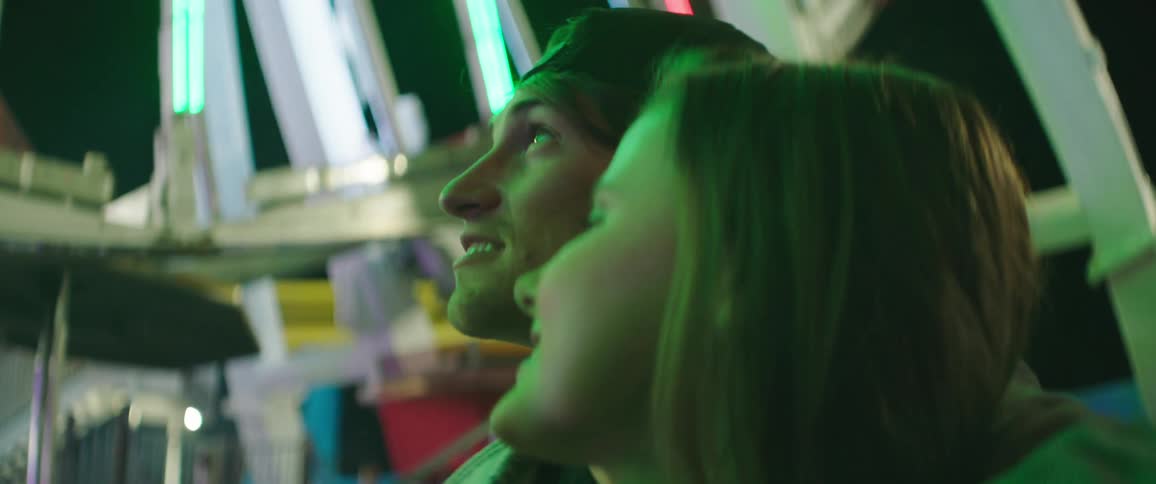 A young man stands with his arm around the shoulders of a young woman as they look up at a colorful ferris wheel