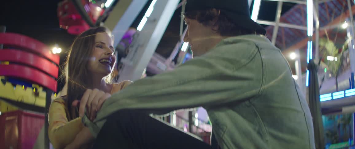 A man and woman laugh and talk while sitting in front of a ride at a carnival