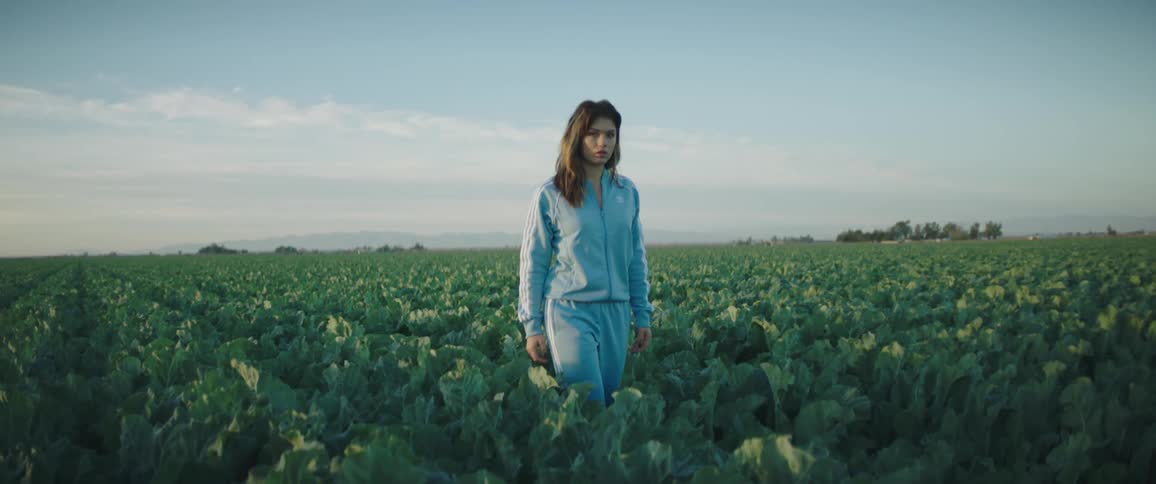 A woman in a track suit walks down a row of crops with a serious look on her face