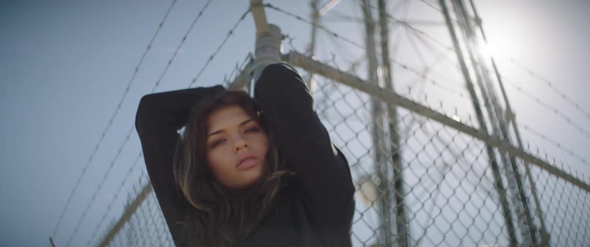 Woman reclined on a wired fence under a transmission tower looking at the camera