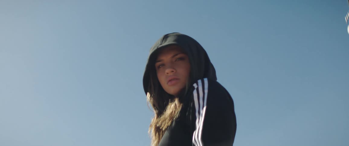 A young woman with the hood of her jacket up stares at the camera while passing by