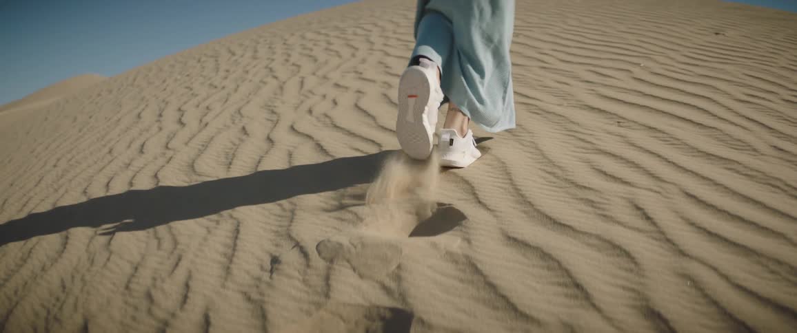 A woman walks up the side of a sand dune in the desert in a dress and sneakers