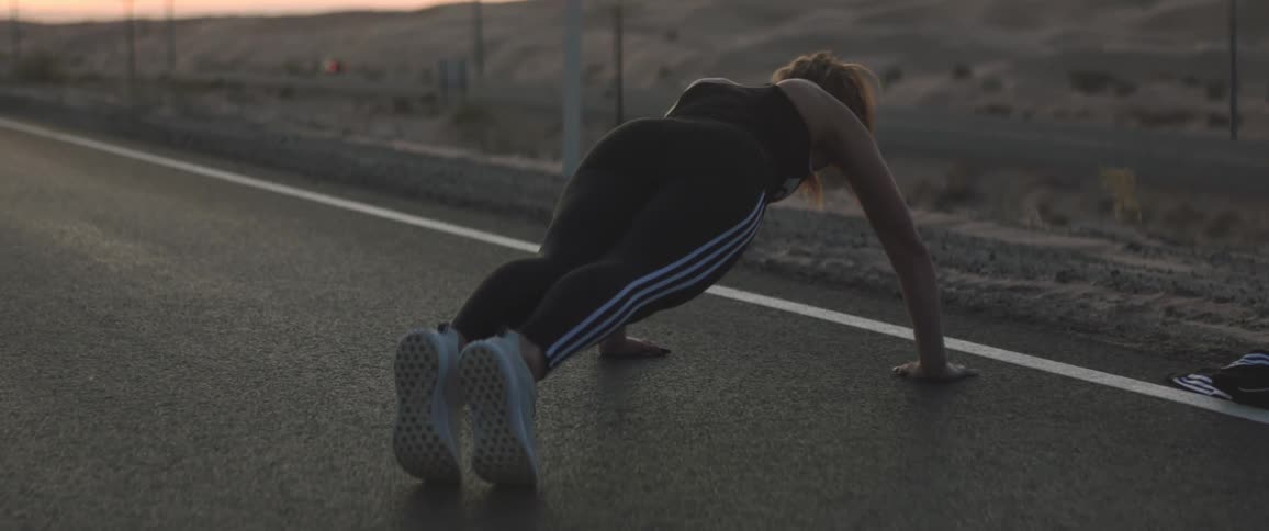 A woman in athletic wears does pushups on a road through a desert in the evening