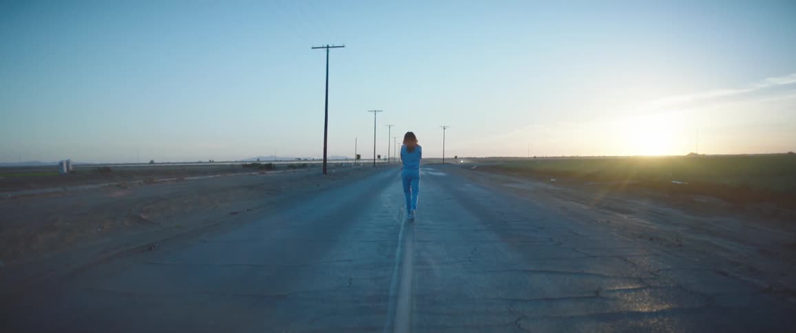 A woman walking on a road next to a grassy field with utility poles the sky and clouds in the background