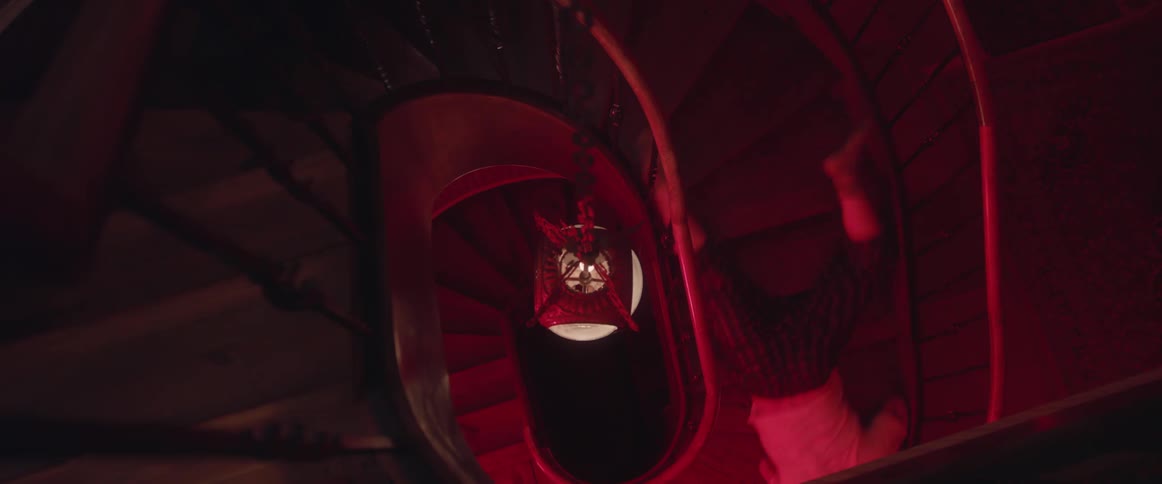 A man with bruises on his legs and ankles falls down a winding staircase as a woman walks slowly behind him