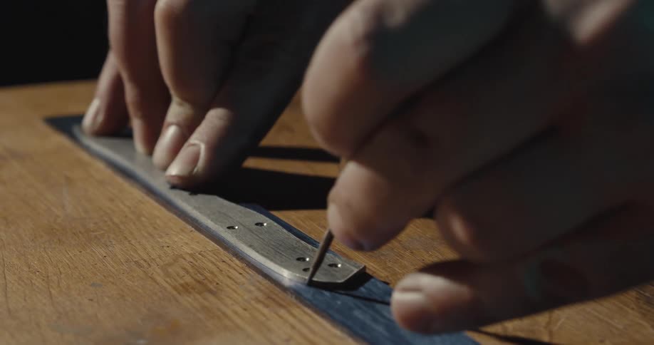 A man uses a sharp tool to draw an outline of a piece of metal on a wood table
