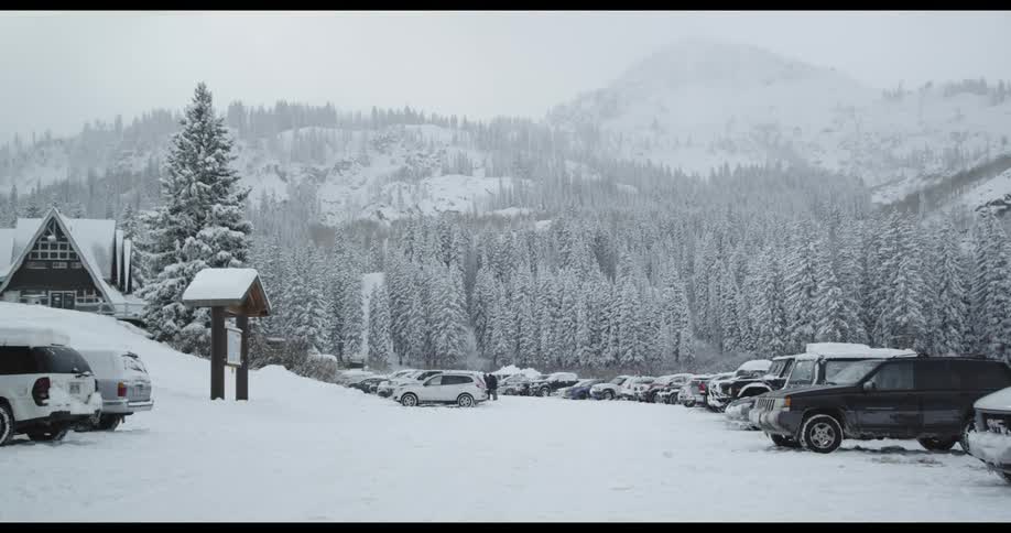 White snow covers a parking lot with parked cars next to a cabin in front of a forest and mountains at daytime