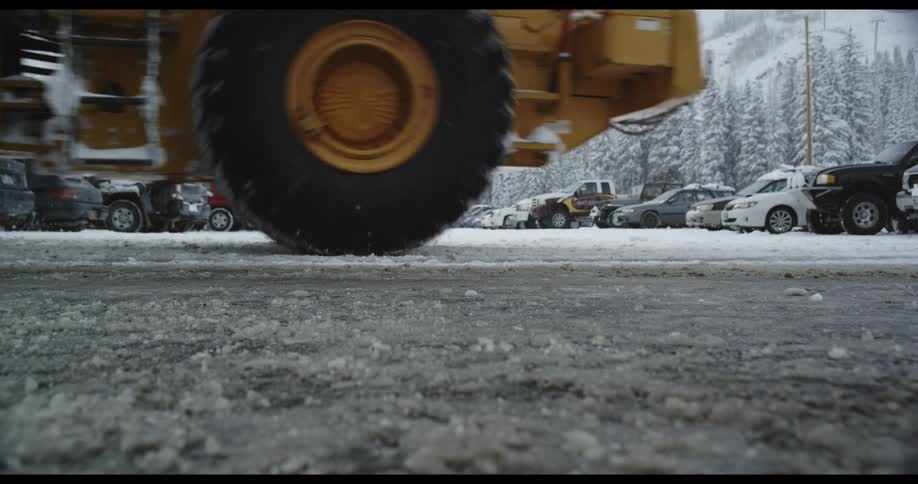 Bulldozer drives through ice covered parking lot full of vehicles backed by conifer forest and mountain slopes