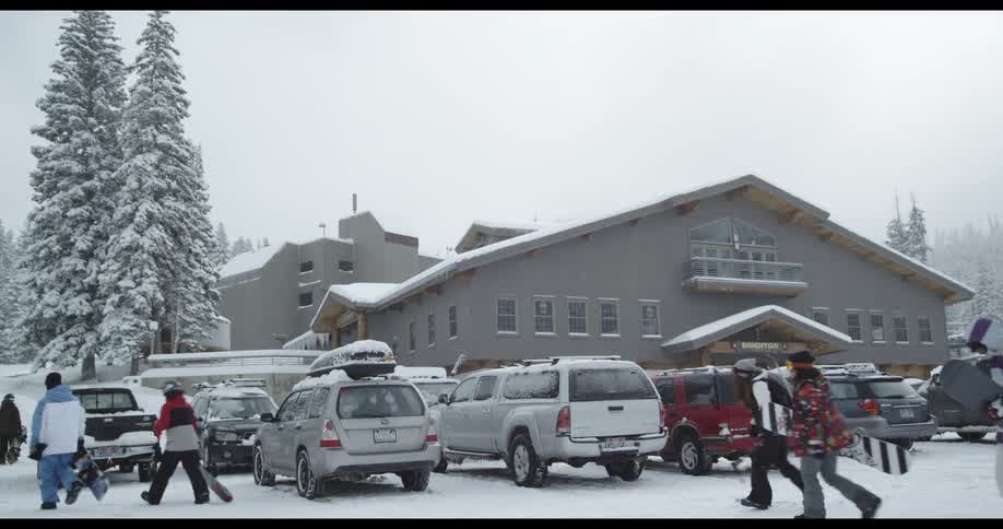 People holding snowboards walking on a snowy day near a building and cars