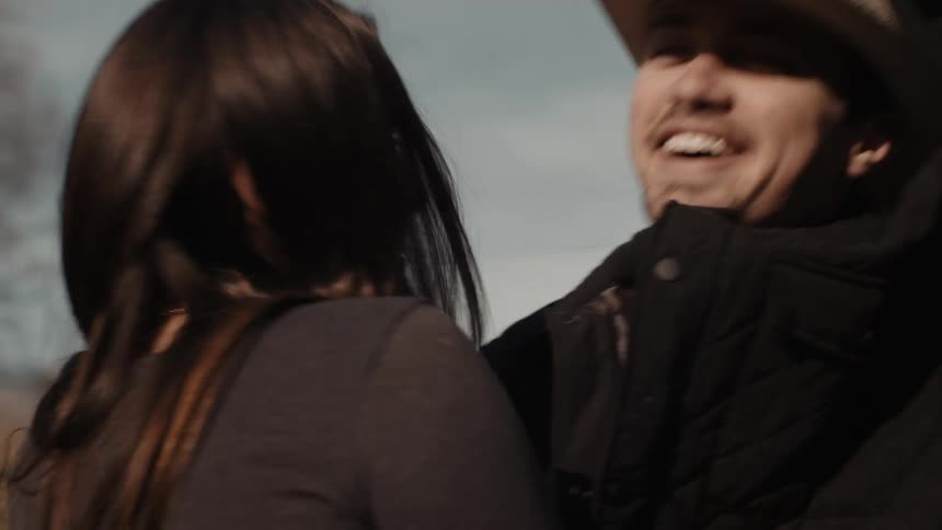 A man holds his hat to his head as a woman embraces him outdoors