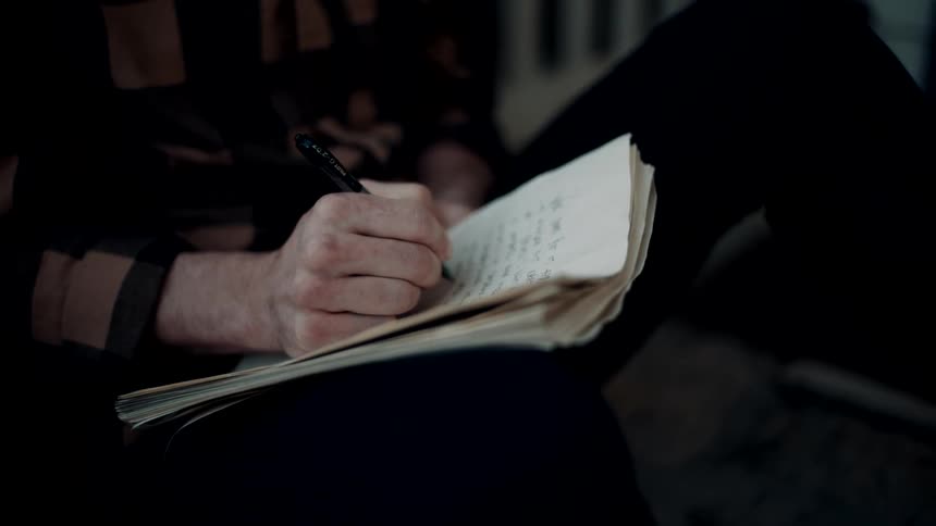 A man is sitting indoors and writing on paper