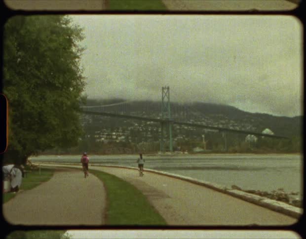 Two cyclists riding bicycles on a road next to a river under a bridge on film