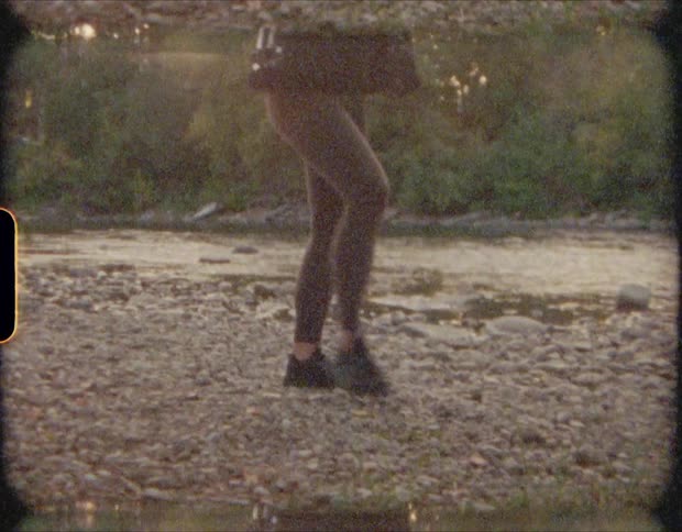 Woman searches through stones on the ground then grabs one and skips it into a river next to her