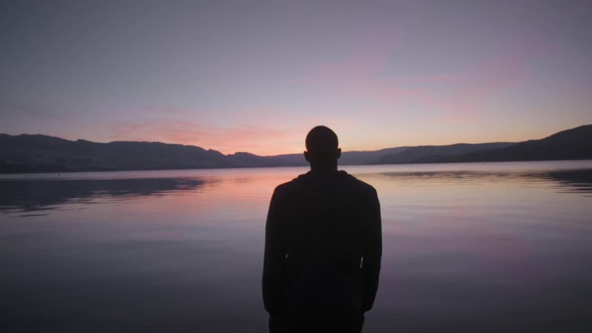 A man standing next to a lake while he watches the sun set behind the mountains