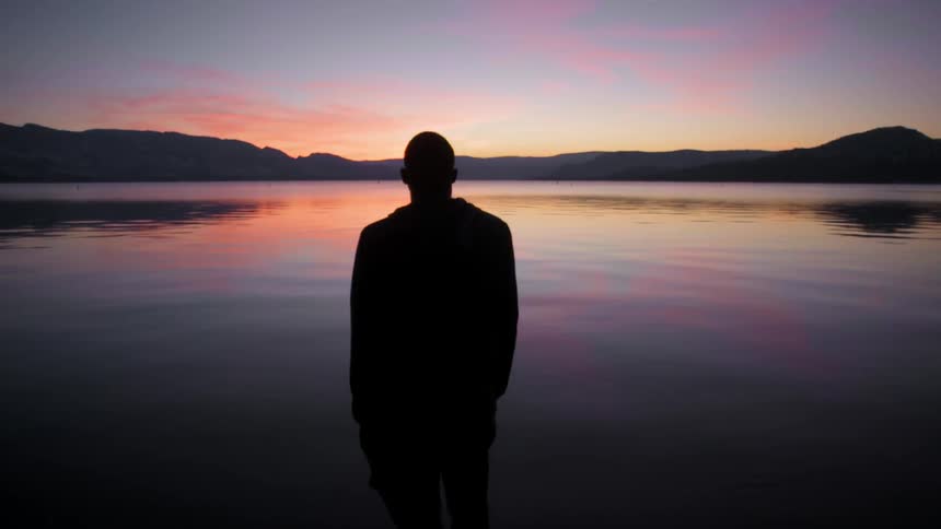 A man stands in the dark before a lake at dawn