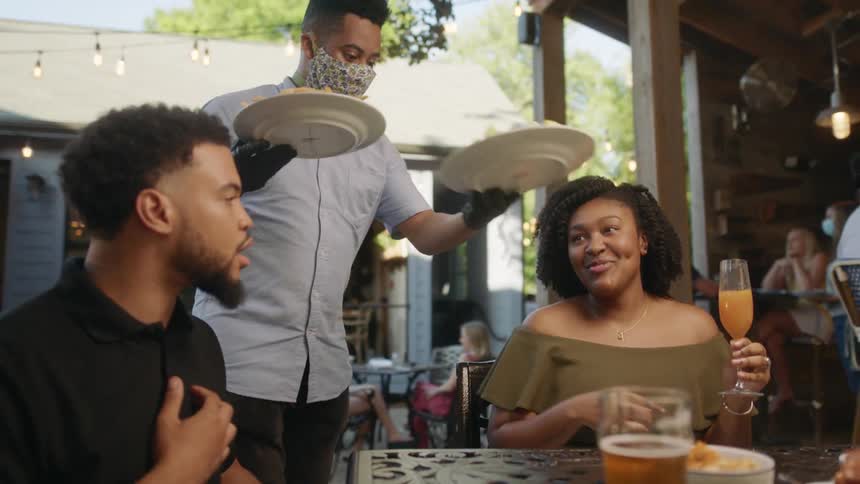 A server in a mask places plates of food before customers sitting at an outdoor table at a restaurant on a sunny day