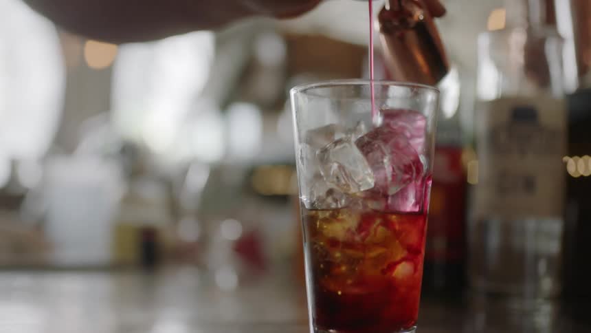 A bartender pours pink liquid into a glass filled with ice cubes and soda sitting on a counter