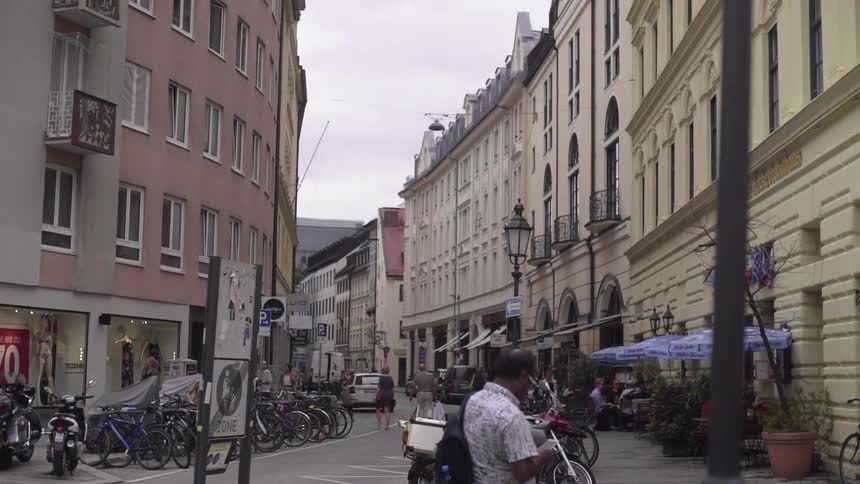 Bicycles and motorcycles are parked near buildings as pedestrians walk around