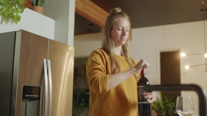 A woman standing in her kitchen holds a bottle of red wine and begins to open it