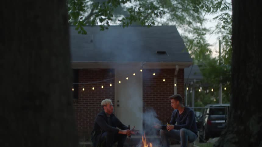 Two male teenagers sit in a backyard around a fire while talking at dusk